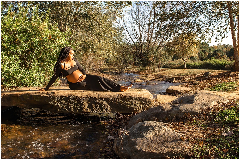 maternity session at Gerrard Landing Park in Roswell, Georgia. Mom is wearing a black dress with the stomach open for viewing. Mom is sitting on a big rock with her hands on her belly